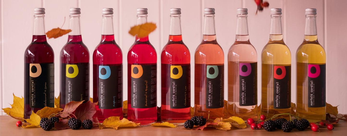 9 x 750ml glass bottles of norfolk cordial placed in a line on a wooden table and white washed wooden panelling behind