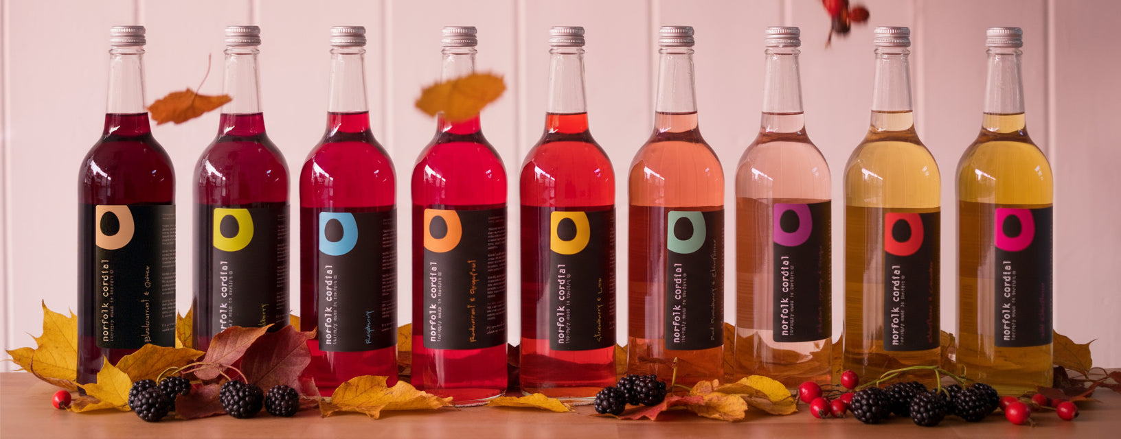 9 x 750ml glass bottles of norfolk cordial placed in a line on a wooden table and white washed wooden panelling behind