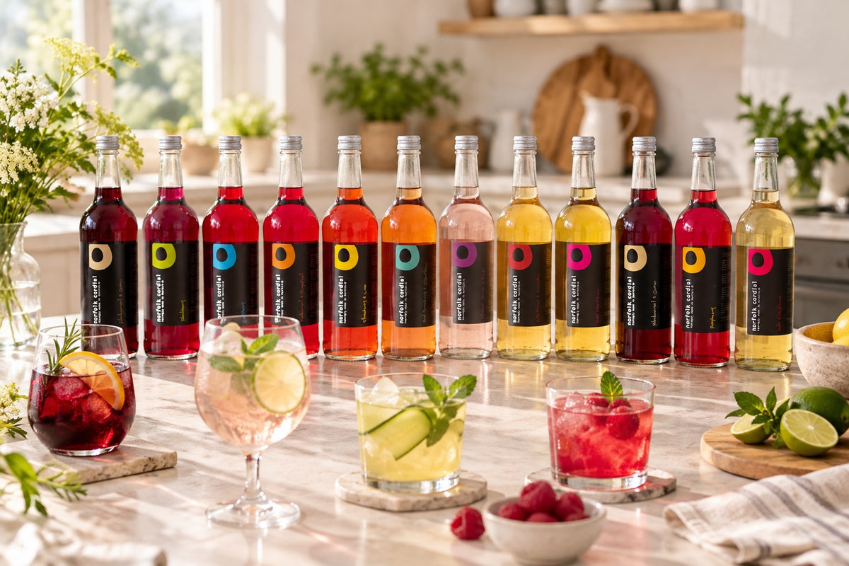 Twelve 750ml Norfolk Cordial bottles displayed in a bright contemporary kitchen with colourful cordial drinks, citrus, mint and fresh raspberries in the foreground.