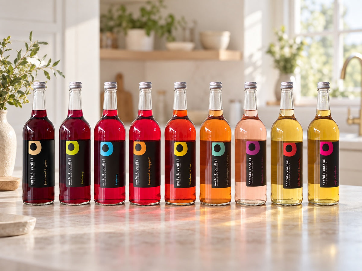 Eight 750ml Norfolk Cordial bottles in assorted flavours arranged on a pale stone worktop in a bright contemporary kitchen.