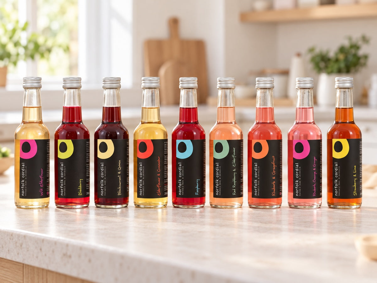 Nine 250ml Norfolk Cordial bottles in assorted flavours displayed on a pale stone worktop in a bright contemporary kitchen.