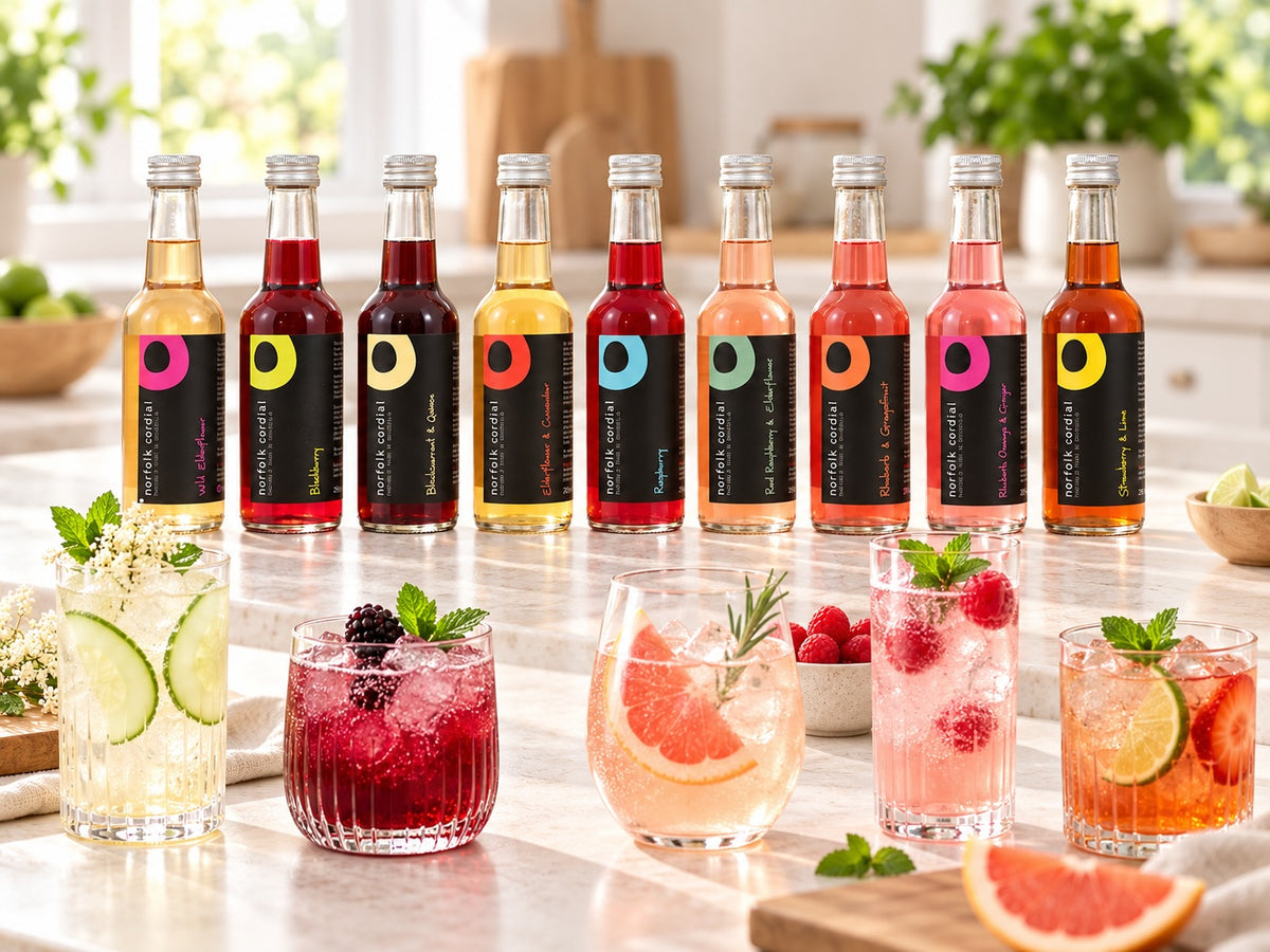 Nine 250ml Norfolk Cordial bottles displayed in a bright contemporary kitchen with colourful cordial drinks, fresh berries, citrus, cucumber and mint.