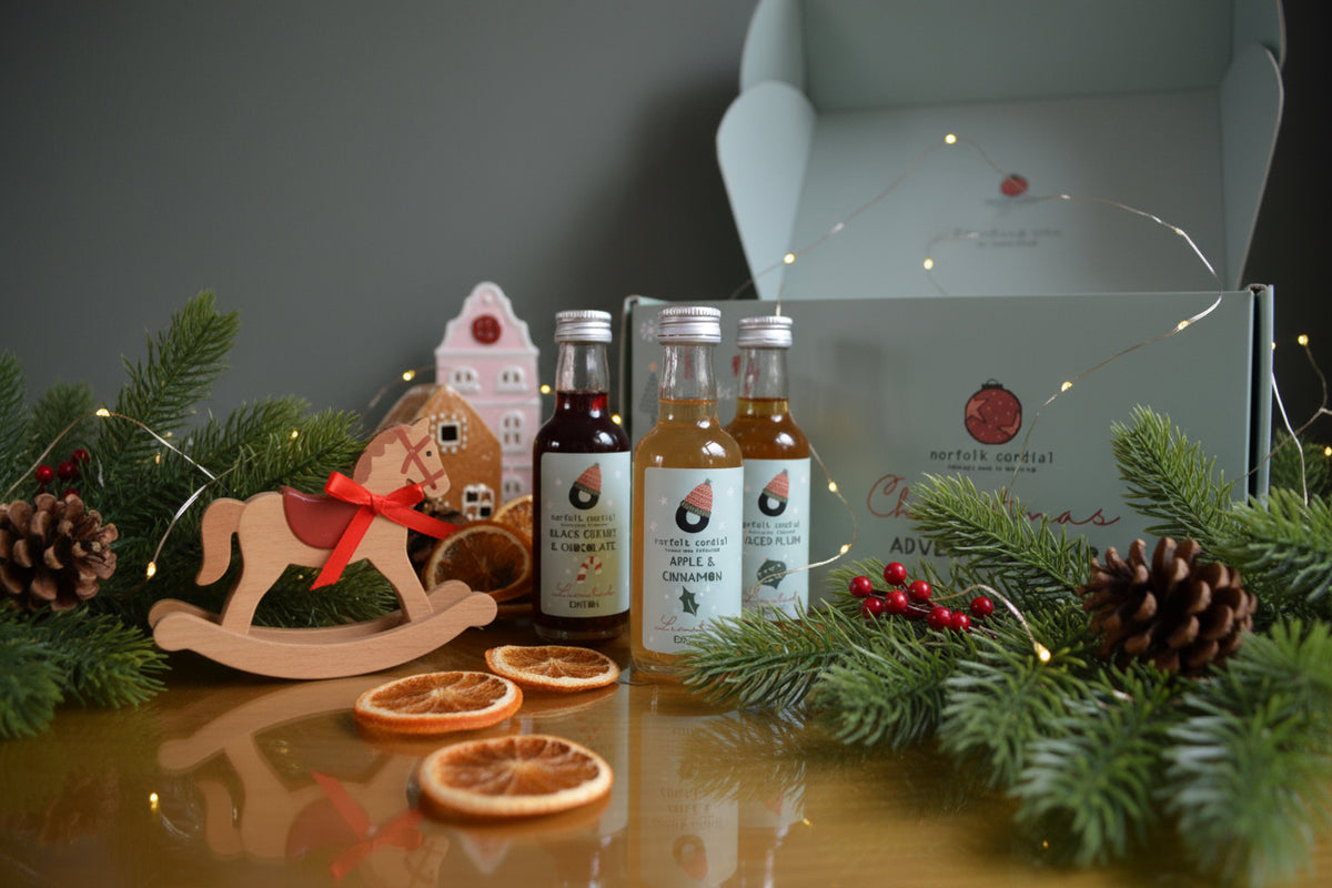 Individual Norfolk Cordial mini bottles displayed in front of an open Christmas advent calendar with festive greenery and dried orange slices.